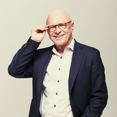 Porträtt av en äldre man i kavaj och glasögon som ler och justerar sina glasögon, fotograferad i studio.